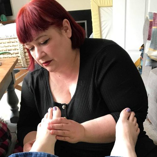 Image of Anita Blizard performing reflexology on some feet with painted toenails. Anita is dressed in black with a red bob hair style.